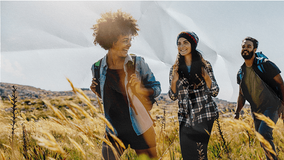 Three young adults joyfully walking through a sunny field of tall grass with hills in the background.