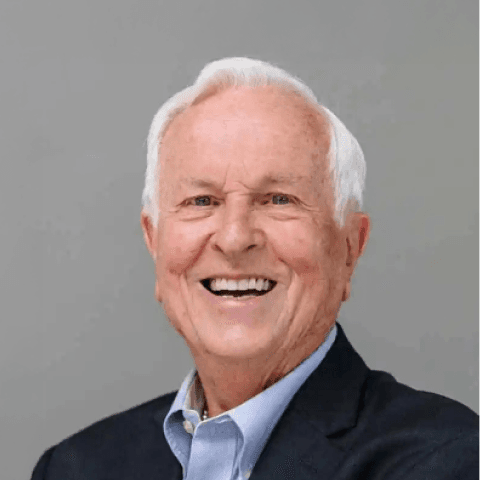 A smiling elderly man with short white hair, wearing a black blazer and blue shirt, against a plain gray background. He exudes warmth and friendliness.