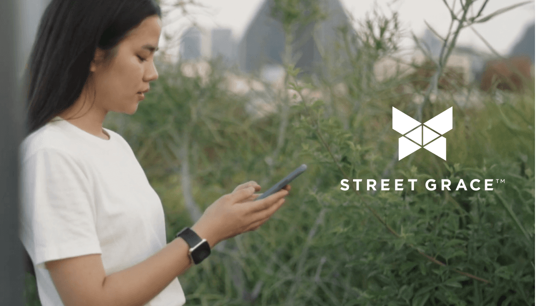 A woman using her phone stands outdoors next to the Street Grace logo.