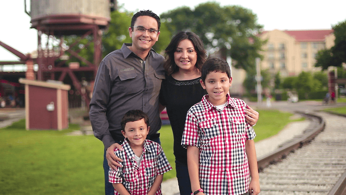 Smiling parents with their two young sons pose beside train tracks in a leafy park.