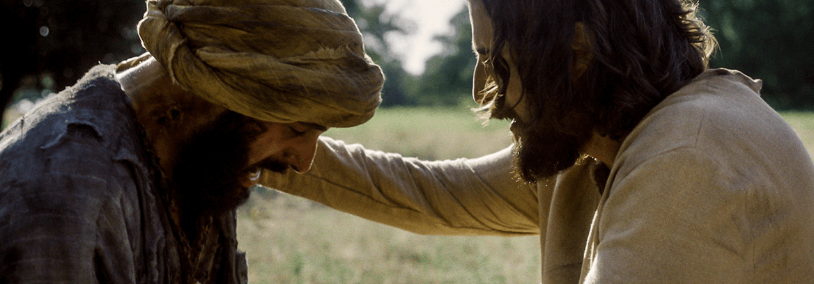 Jesus comforts a distressed man wearing a headwrap in an open field.