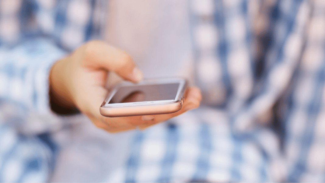 Hand holding a smartphone with the person wearing a blue plaid shirt in the background.