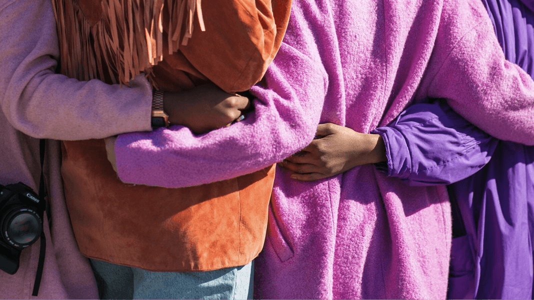 Group of people wearing purple, pink, and rust-colored clothing stand arm in arm.