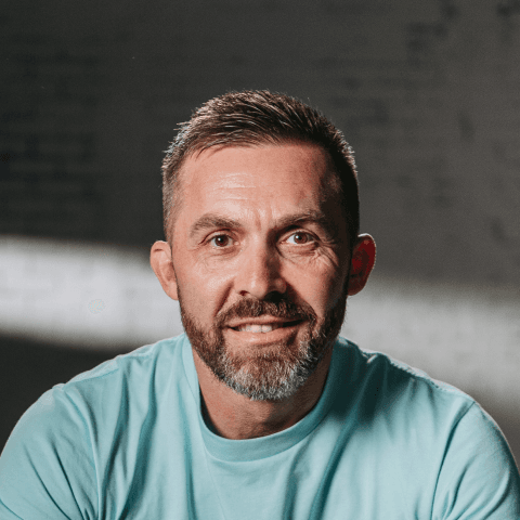 Bearded man in a light blue shirt smiles warmly against a softly lit, dark background. The image conveys a friendly and approachable tone.