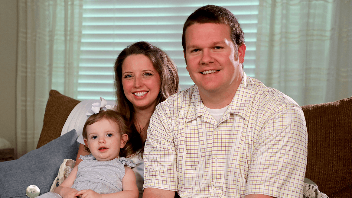 A smiling couple with a young child sit together on a couch in a warmly lit room.