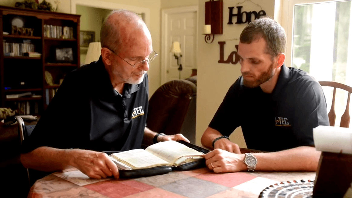 Two men sit at a table in a cozy living room, reading and discussing a Bible together.