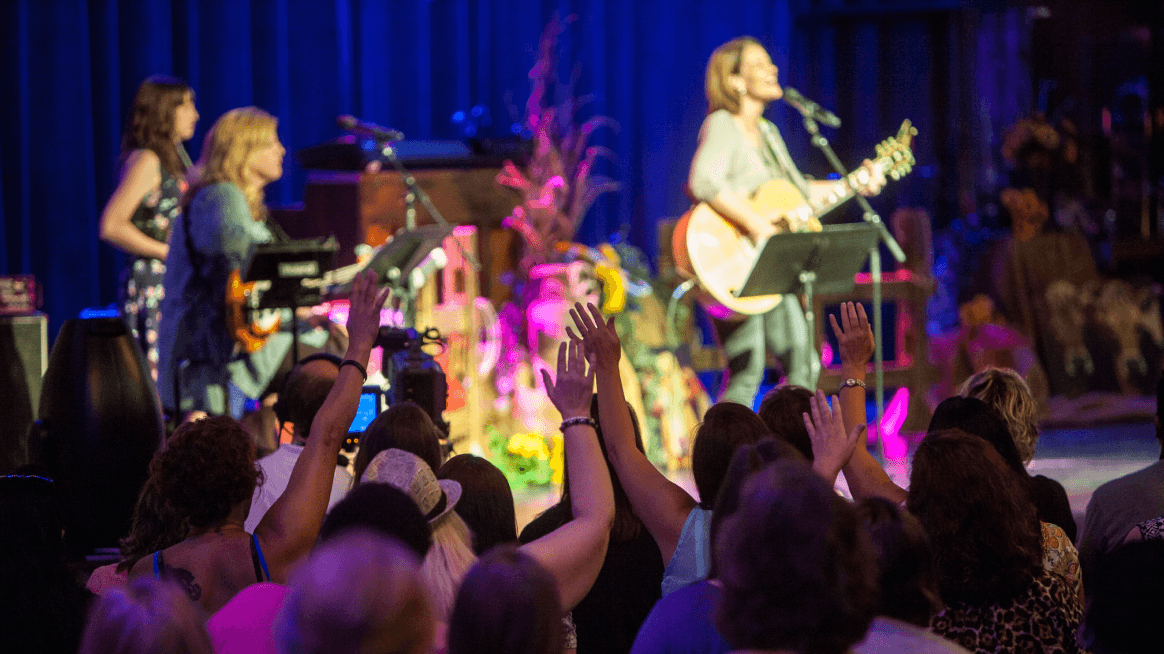 Worship team of women singing and playing instruments on stage during a church service.