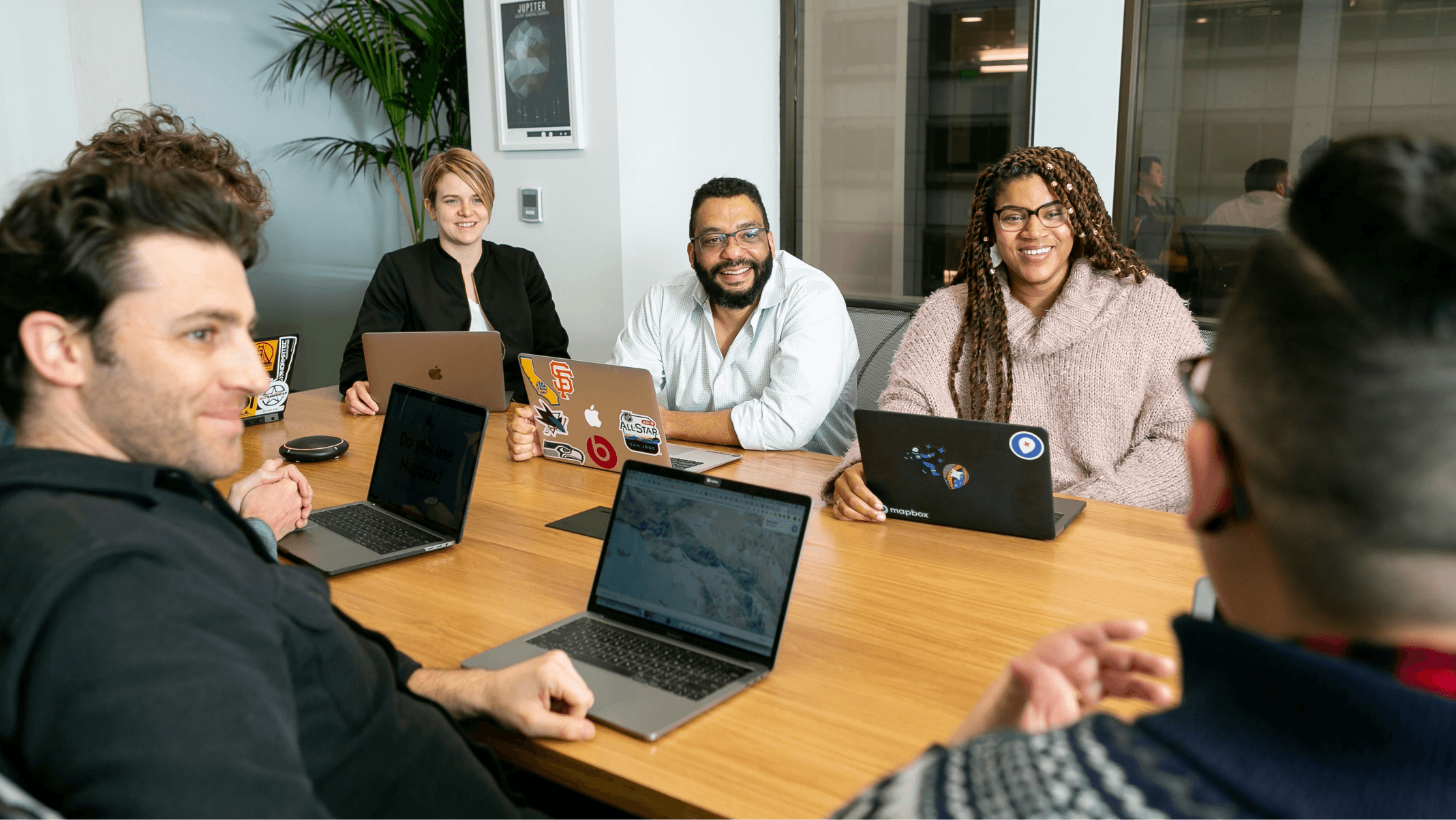 Team of six people sitting around a conference table with laptops, smiling and discussing a project.