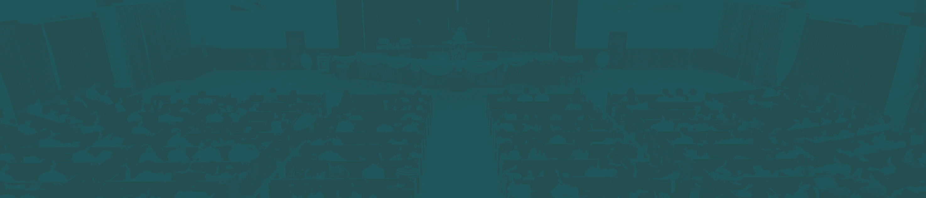 Wide-angle photo of a church sanctuary filled with seated attendees facing a stage with musical instruments and a speaker at the podium, overlaid with a dark teal tint.